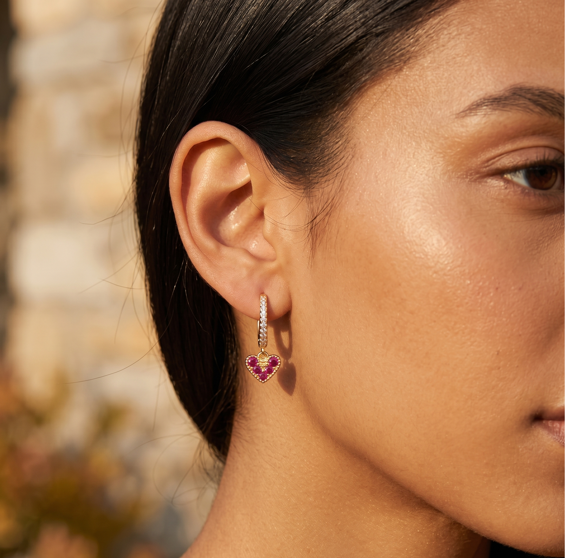 Close-up of a woman wearing a heart-shaped earring with pink gemstones against a blurred natural background.