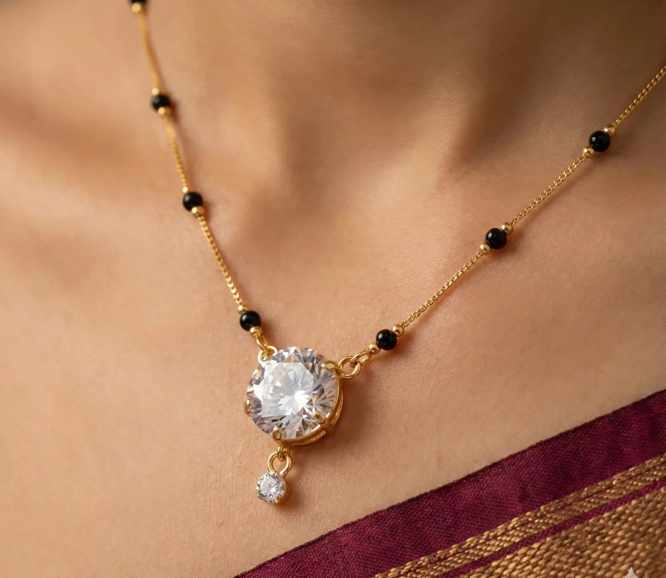 Gold necklace with a large central gemstone and smaller pendant on a person wearing a maroon saree.