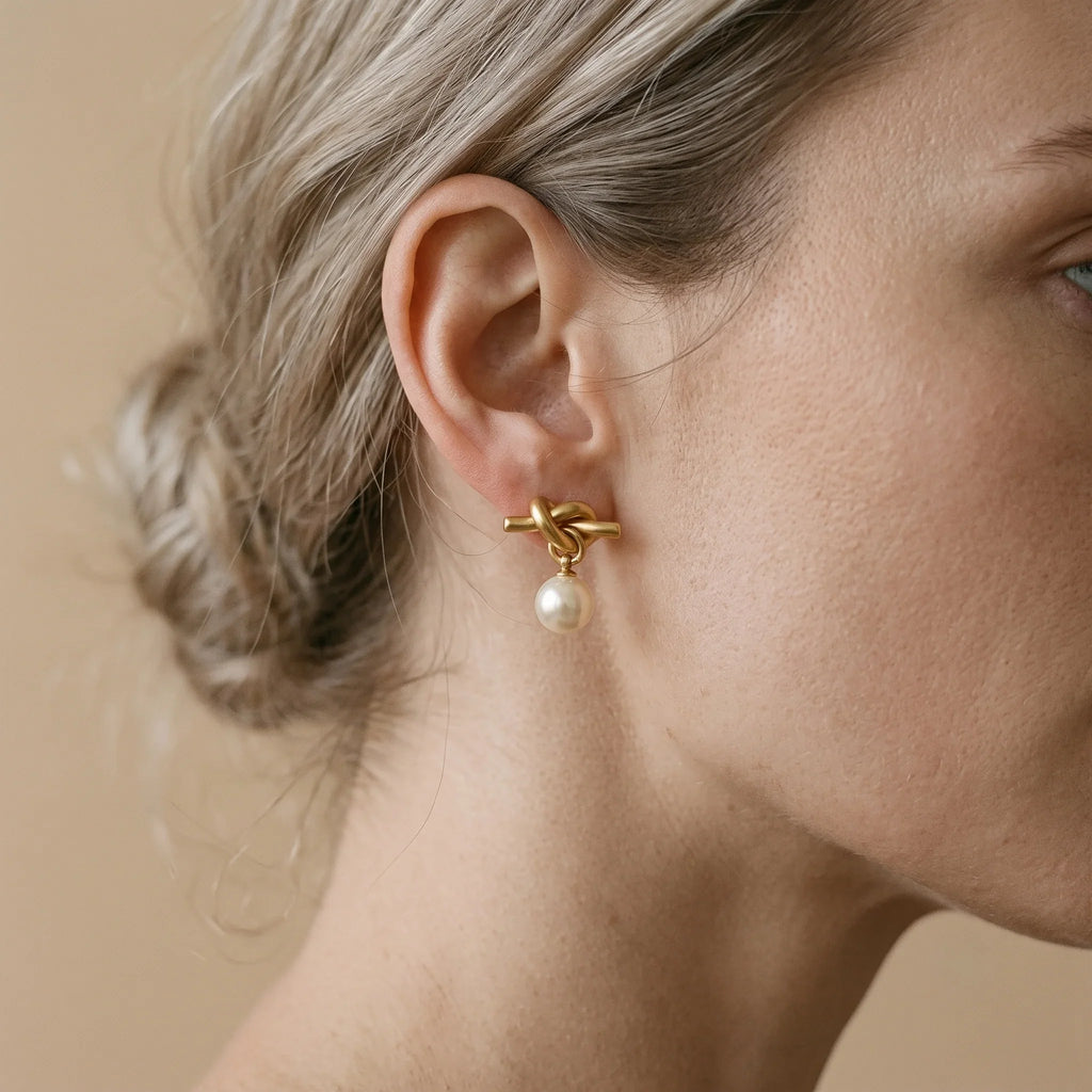 A close-up of a woman's ear wearing a gold earring with a twisted knot design and a small dangling pearl, against a soft beige background.