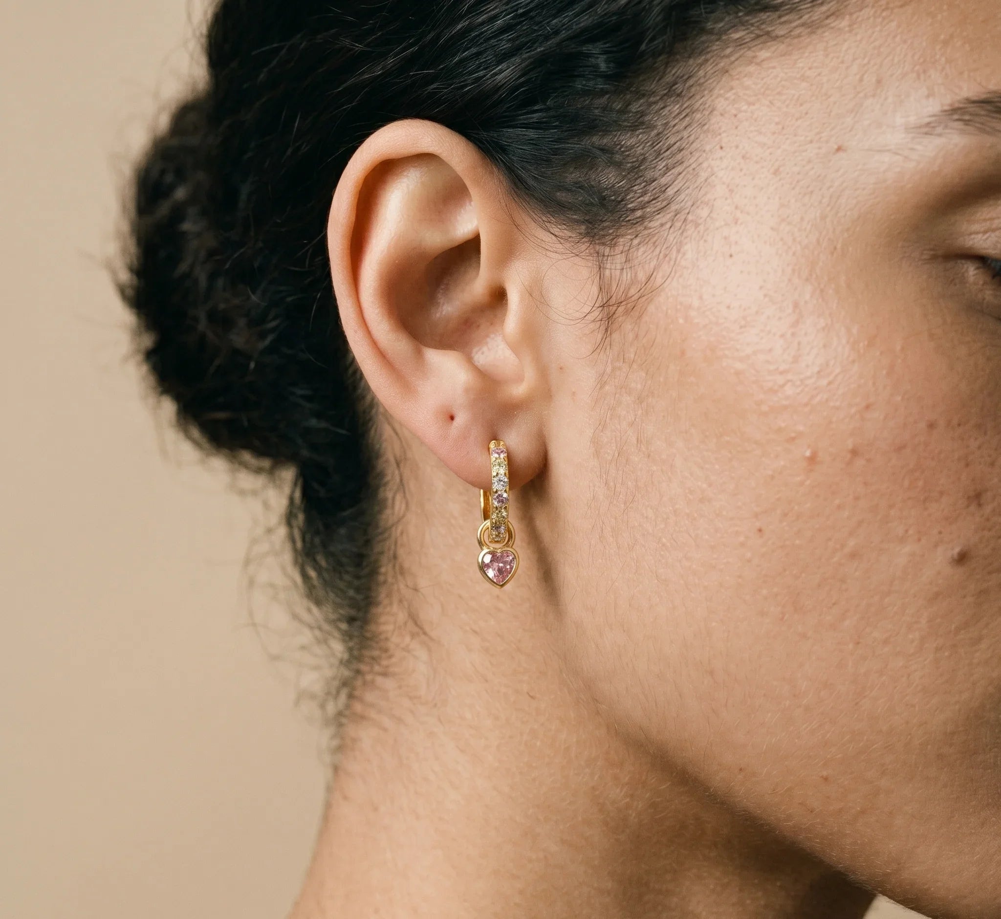 Close-up of an ear wearing a gold earring with a pink gemstone on a beige background.