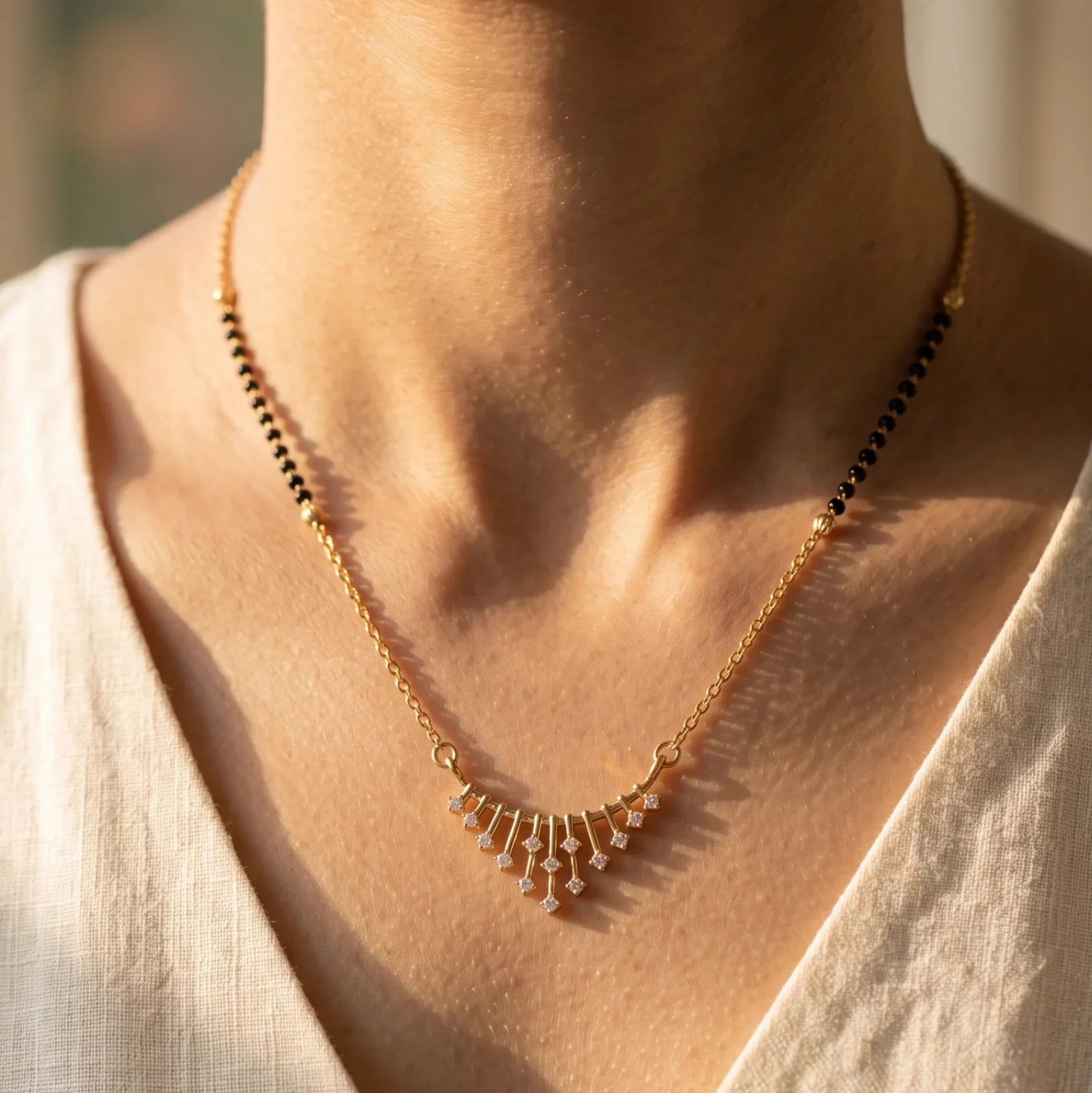 A close-up lifestyle shot of a model wearing a delicate gold necklace with a fan-shaped cluster of small clear stones, styled with a cream-colored linen top.