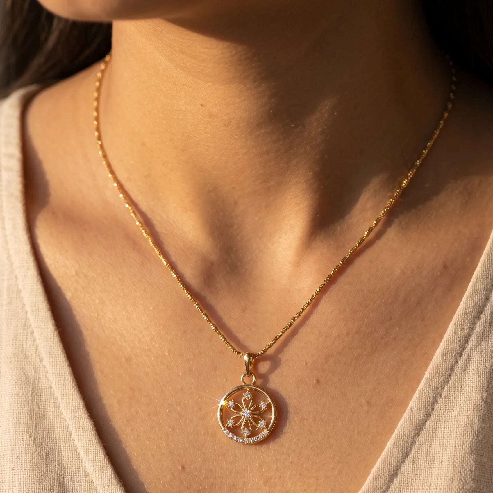 A close-up of a person wearing a delicate gold chain necklace with a circular pendant featuring a floral starburst design set with small clear stones, styled with a beige linen V-neck top.