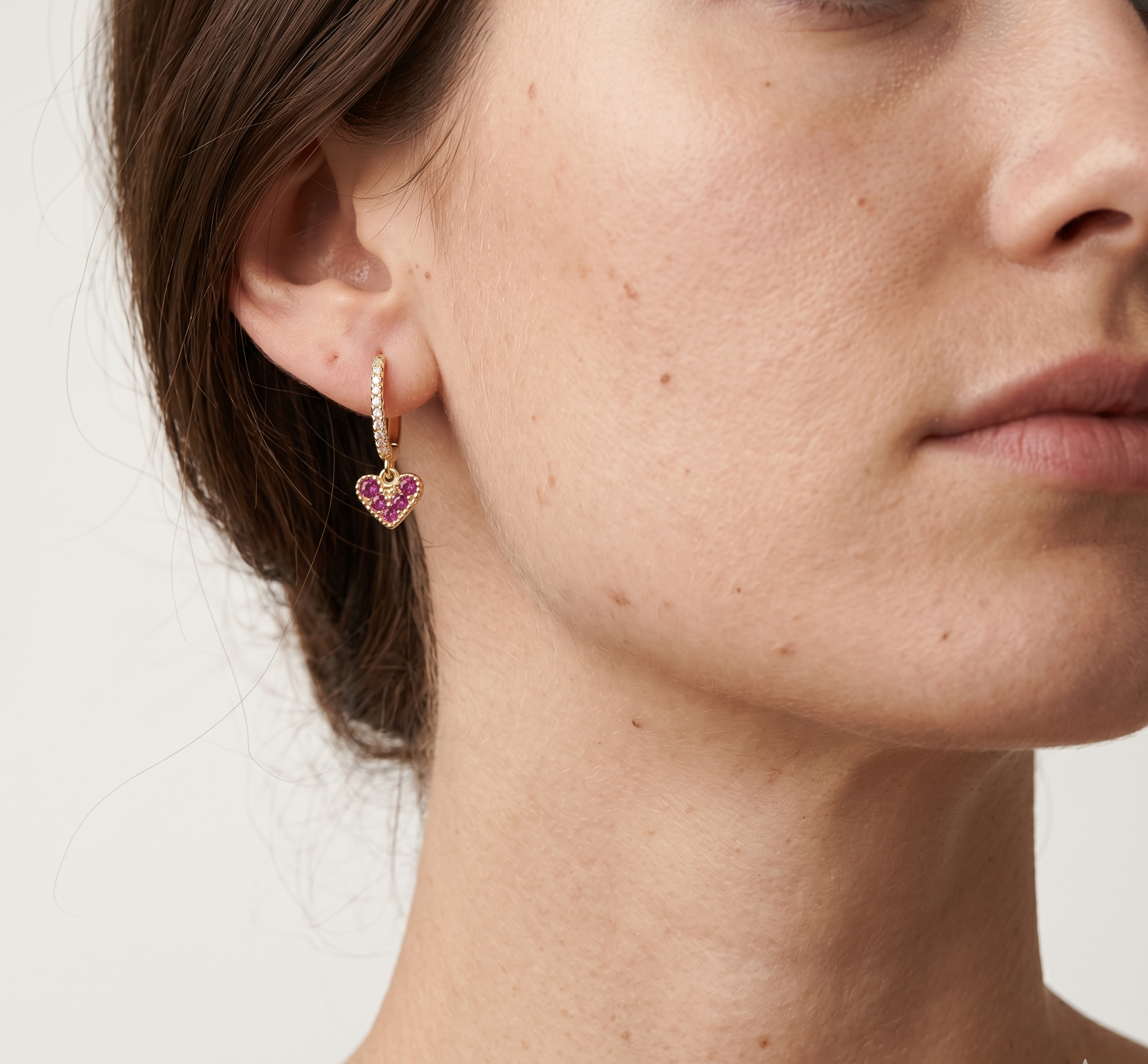 Close-up of a woman wearing gold heart-shaped earrings with pink stones on a neutral background