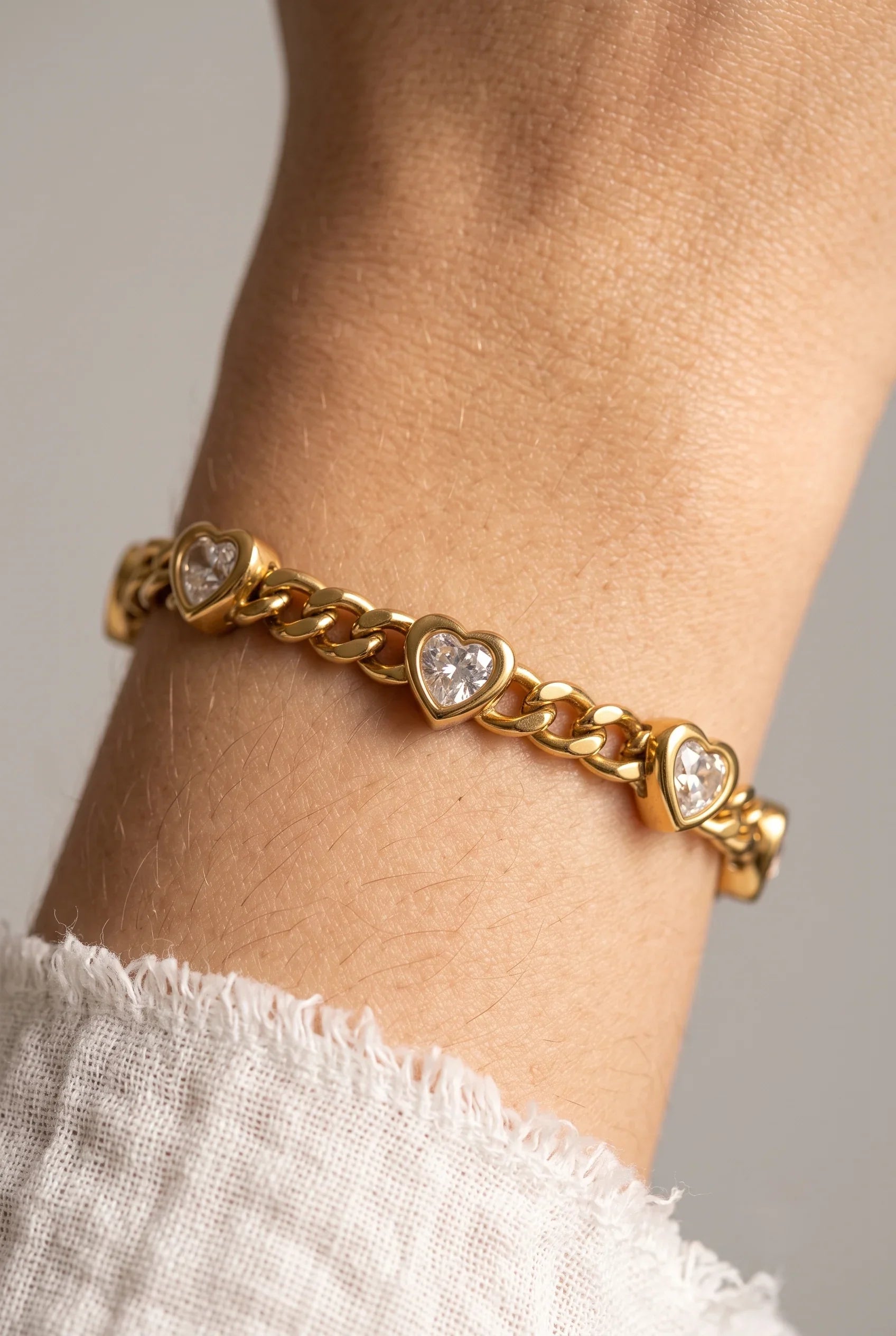 A gold chain bracelet with three heart-shaped clear gemstone accents, shown worn on a person's wrist against a soft gray background.