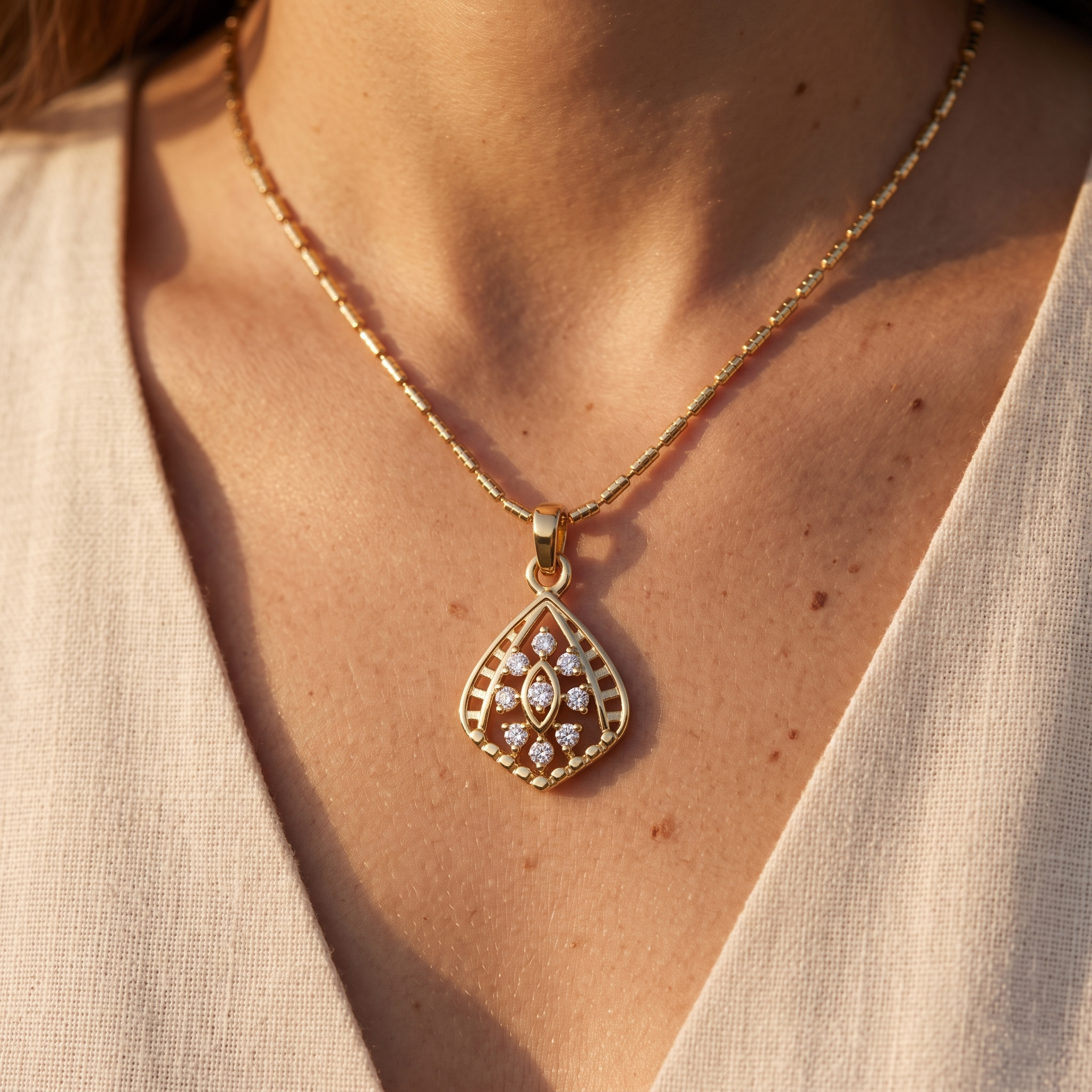 A close-up lifestyle shot of a gold teardrop pendant necklace with a diamond-encrusted center, worn by a model in a beige linen top.