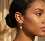 Close-up of a woman wearing a gold earring with a clear gemstone against a neutral background
