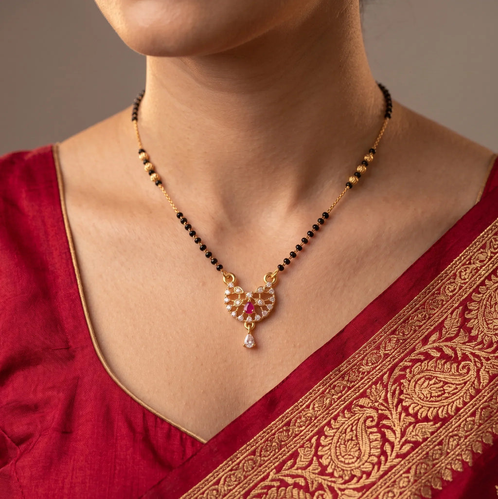 A close-up of a woman wearing a gold and black beaded necklace with a detailed gold pendant featuring red and white stones, paired with a red embroidered blouse.