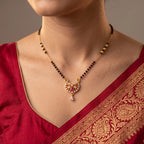 A close-up of a woman wearing a gold and black beaded necklace with a detailed gold pendant featuring red and white stones, paired with a red embroidered blouse.