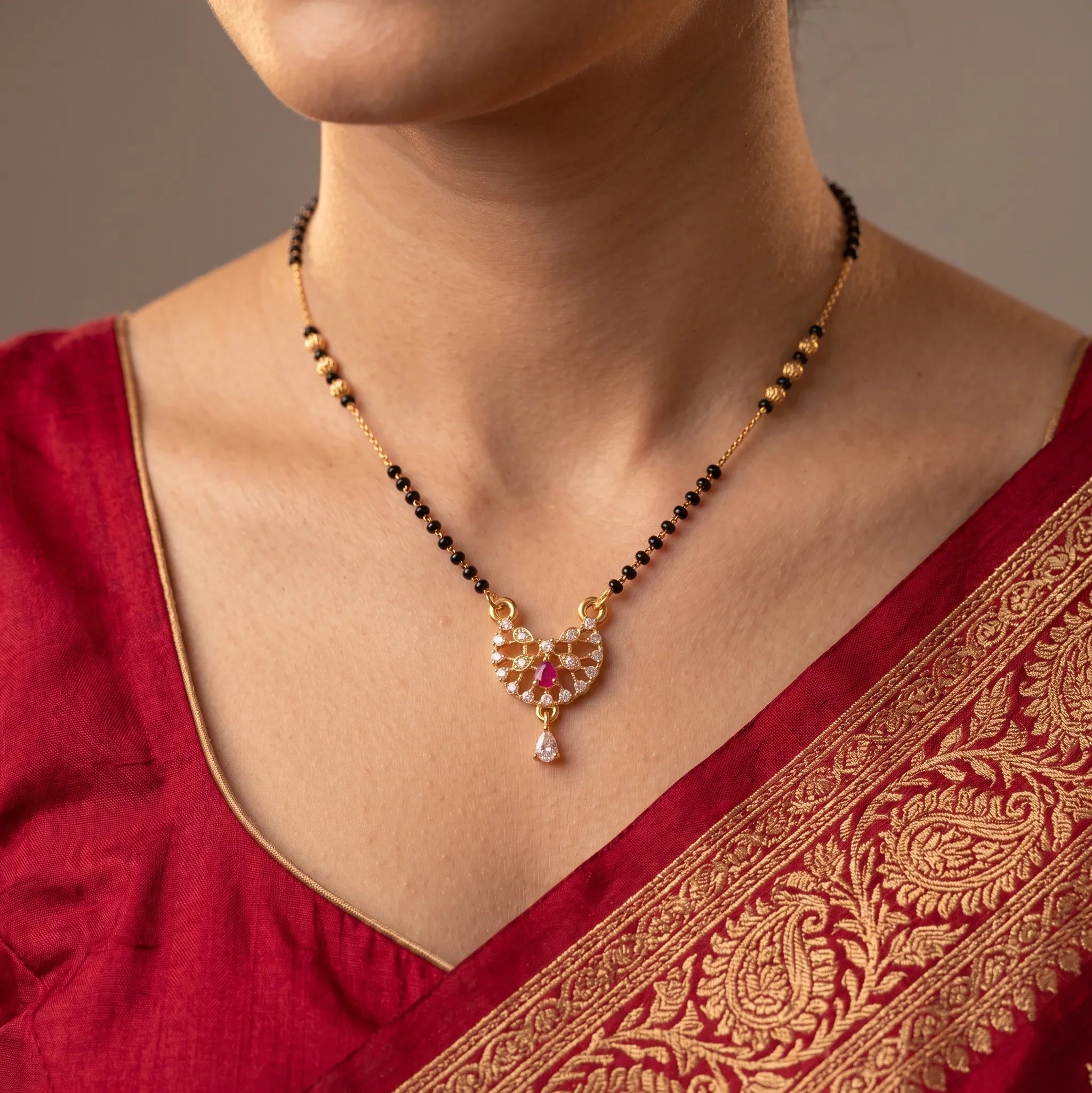 A close-up of a woman wearing a gold and black beaded necklace with a detailed gold pendant featuring red and white stones, paired with a red embroidered blouse.