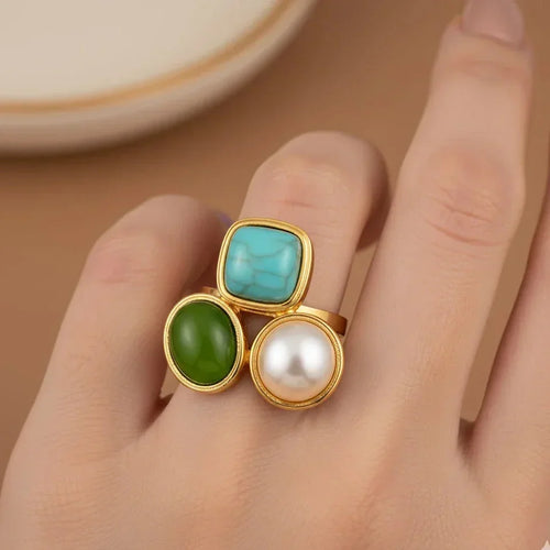 Gold ring with turquoise, green, and pearl stones on a hand against a beige background