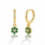 A pair of gold huggie hoop earrings with dangling flower charms. Each flower charm features green gemstones surrounding a small clear center stone, set against a clean white background with a soft reflection below.