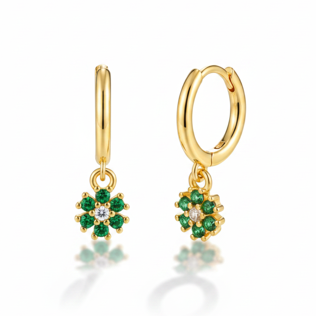 A pair of gold huggie hoop earrings with dangling flower charms. Each flower charm features green gemstones surrounding a small clear center stone, set against a clean white background with a soft reflection below.