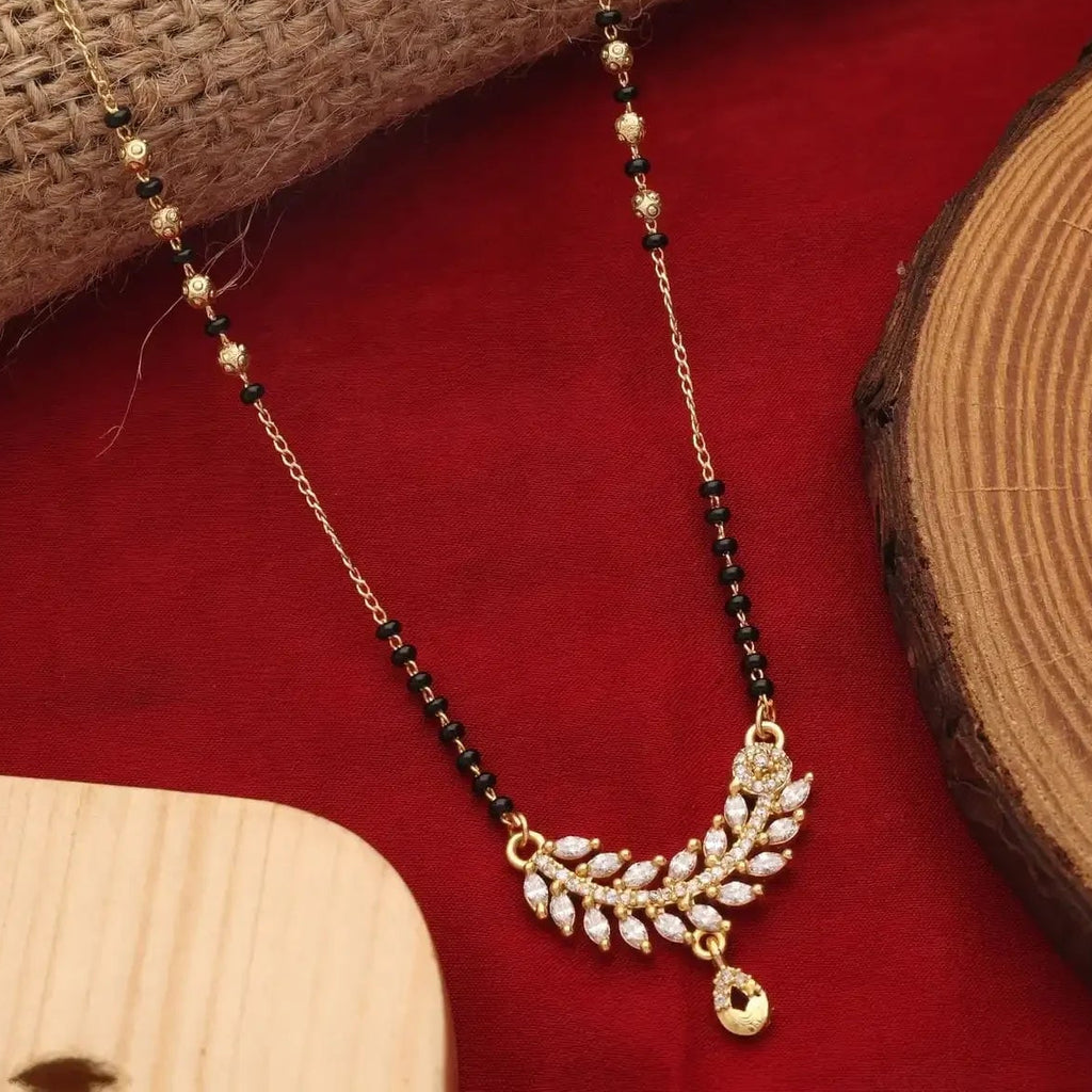 A gold and black beaded necklace with a leaf-shaped crystal pendant, displayed on a red velvet surface with decorative wood and burlap props.